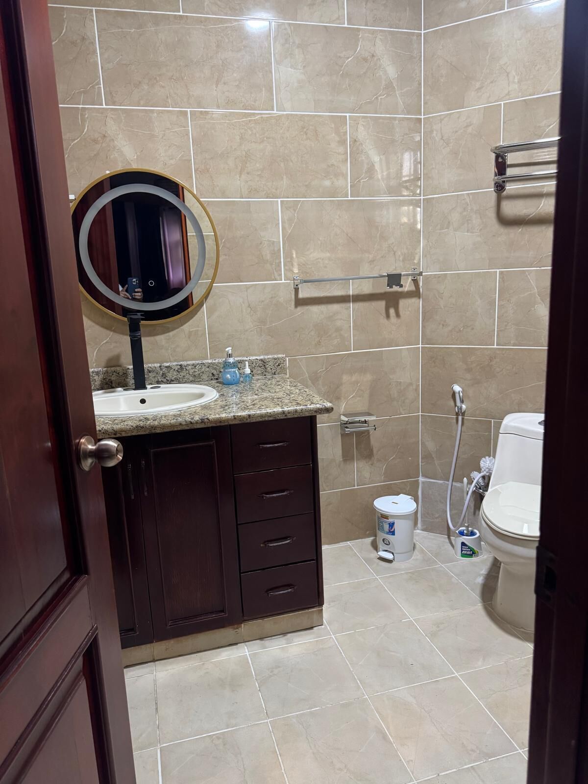A bathroom with a marble-tiled wall, a sink with a round mirror, a toilet, and a shower area.