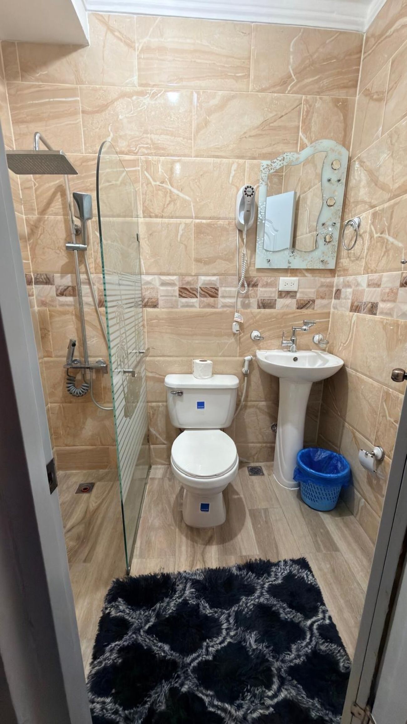 A bathroom with a glass shower enclosure, a toilet, a sink, and a blue trash bin.