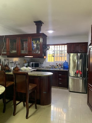 A kitchen with wooden cabinets, a stainless steel refrigerator, a microwave, and a dining area with chairs and a table. - Estudio Carnavalesco (La Vega)