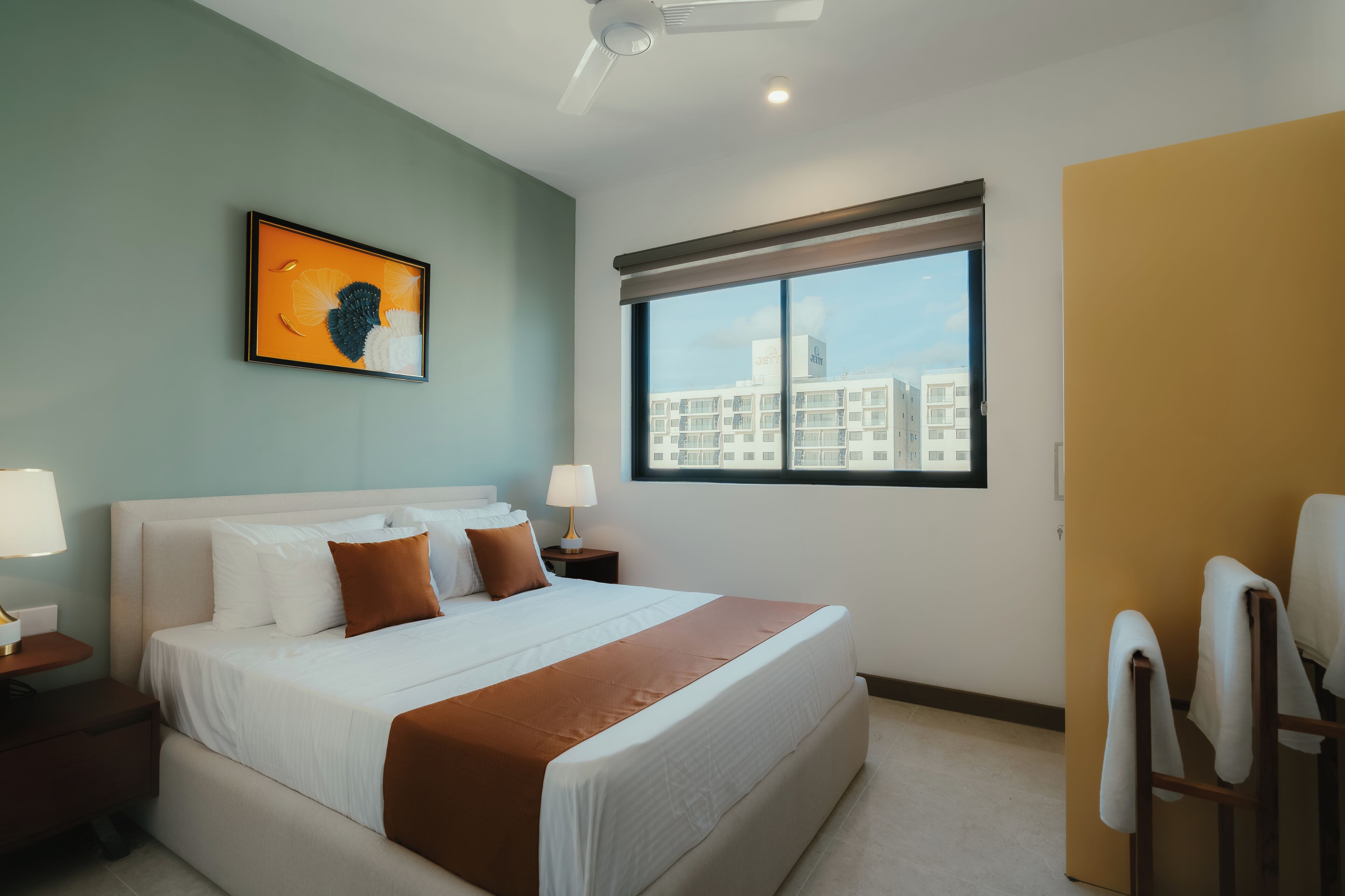 A hotel room with a bed, two bedside tables with lamps, a window with a city view, and a ceiling fan.