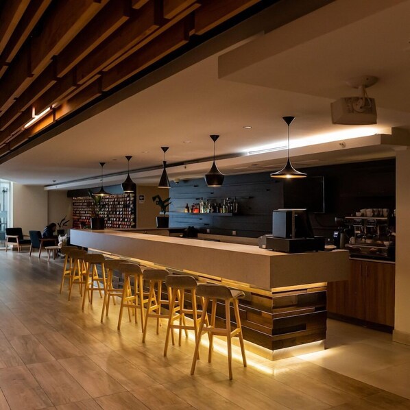 Un área de bar moderna con taburetes de madera, iluminación colgante y una barra con una máquina de café.