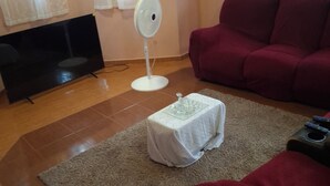 Ein Wohnzimmer mit einem Flachbildfernseher, einem Standventilator, einem roten Sofa und einem kleinen Tisch mit einer Glasplatte.