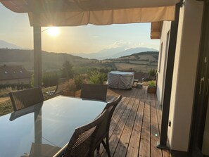 Eine Holzterrasse mit einem Glastisch und Korbstühlen, mit Blick auf ein Tal und Berge in der Ferne.