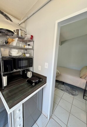 Eine kompakte Küchenzeile mit einer Mikrowelle, einem Toaster und einer Kaffeemaschine auf einem Regal, einem kleinen Schrank darunter und einem Bett im angrenzenden Zimmer.