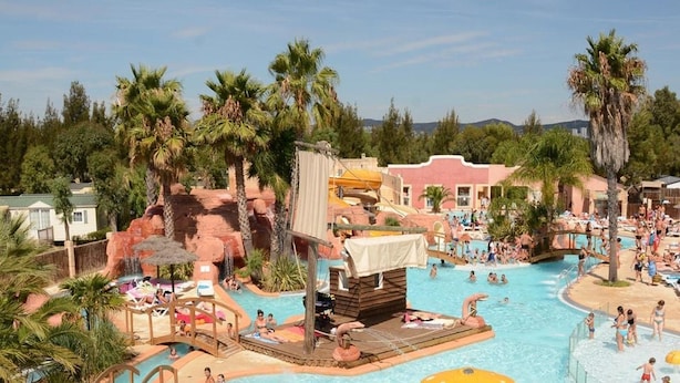 Área de piscina en un resort con múltiples fuentes, palmeras y montañas al fondo.