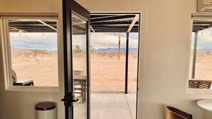 A room with a door leading to a covered outdoor area, a trash bin, a chair, and a welcome mat. - DILIM - Designer Desert Retreat (Homestead Valley)