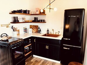 Une cuisine avec des meubles de couleur noire, une cuisinière noire, un réfrigérateur noir, des étagères en bois et une machine à café.
