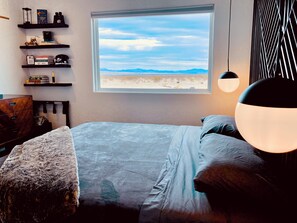 Une chambre avec une grande fenêtre, un lit, un bureau, des étagères remplies de livres et d’objets, et un luminaire suspendu.
