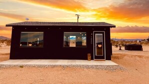 A small, modern building with a dark exterior, large windows, and a door, situated in a desert landscape with a sunset in the background. - DILIM - Designer Desert Retreat (Homestead Valley)