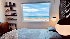 A bedroom with a bed, a window with a view of a desert landscape, a shelf with books and decorative items, and a hanging light fixture. - DILIM - Designer Desert Retreat (Homestead Valley)