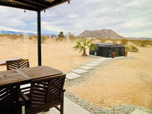 Un espace extérieur aménagé avec une table et des chaises, un palmier et un bâtiment, le tout situé dans un paysage désertique avec des montagnes en arrière-plan.