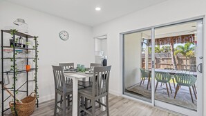 Un espace repas comprenant une table, des chaises, une horloge et une porte-fenêtre coulissante donnant sur une terrasse extérieure.