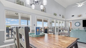A dining area with a wooden table and chairs, a chandelier, and a view of the outdoors through large windows. - Catch & Release ~ Bay Front ~ Sleeps 12 (Dauphin Island)