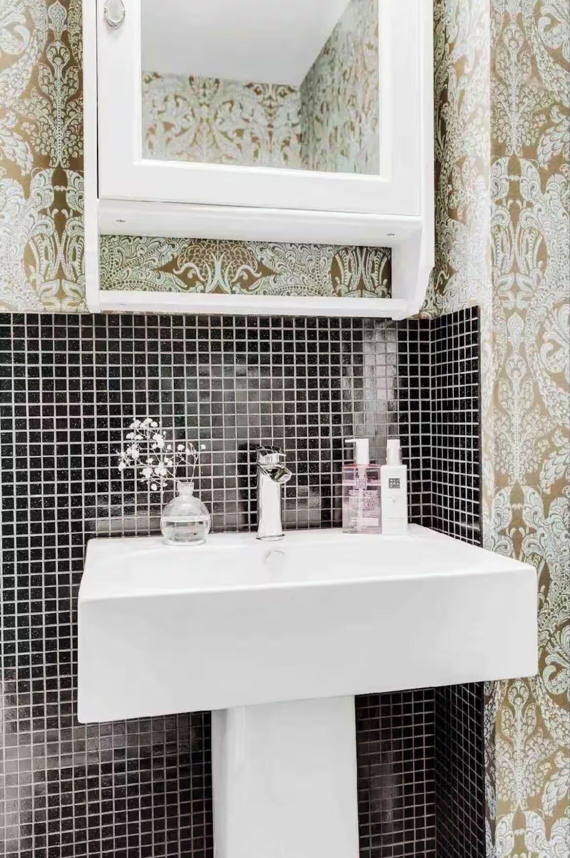 Een badkamer met een wit wastafelmeubel, een spiegel erboven en behang met een patroon.