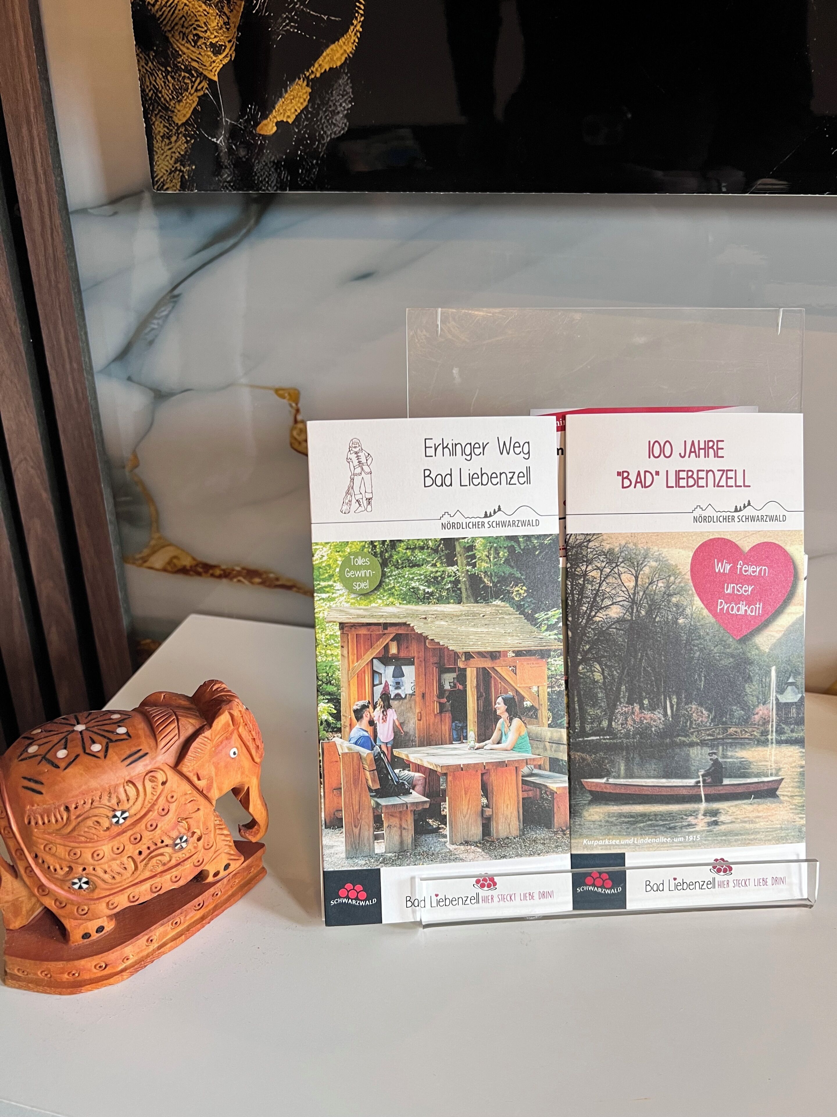 Dos portadas de libros tituladas 'Erkinger Weg Bad Liebenzell' y '100 Jahre 'Bad' Liebenzell', con una escultura de elefante de madera y un fondo de mármol.