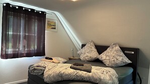 Et soveværelse med seng, puder, dyne, et natbord, en lampe og et vindue med gardiner.