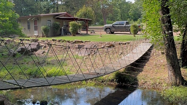 A wooden bridge over a small stream, surrounded by trees and a house in the background.