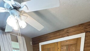 A bedroom with a large bed, a ceiling fan, wooden panel walls, a window with curtains, and a bedside lamp.