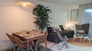 A dining area with a wooden table and chairs, a potted plant, a bed with a green bedspread, a chair with a furry throw, and a mirror on the wall. - Charming gîte de la Cour Suard (Cigogné)