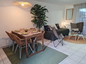 A dining area with a wooden table and chairs, a potted plant, a bed with a green bedspread, a chair with a furry throw, and a mirror on the wall. - Charming gîte de la Cour Suard (Cigogné)
