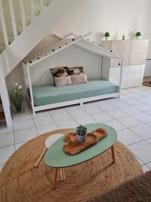 A room with a white staircase, a green sofa bed with pillows, a small table with a plant, and a woven rug. - Charming gîte de la Cour Suard (Cigogné)
