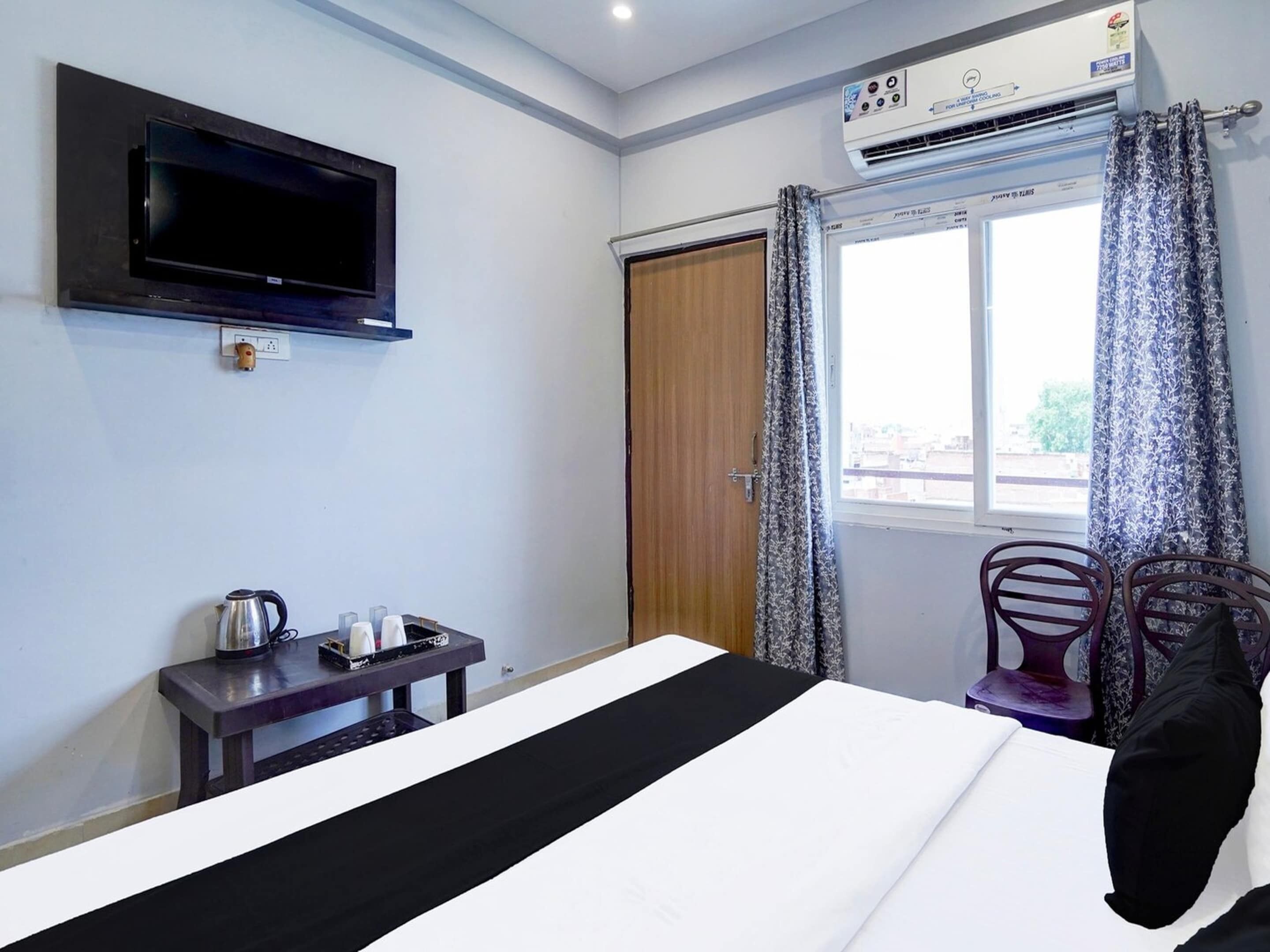 Quarto de hotel com cama, televisão fixada na parede, mesa pequena com chaleira e xícaras, janela com cortinas e aparelho de ar condicionado.