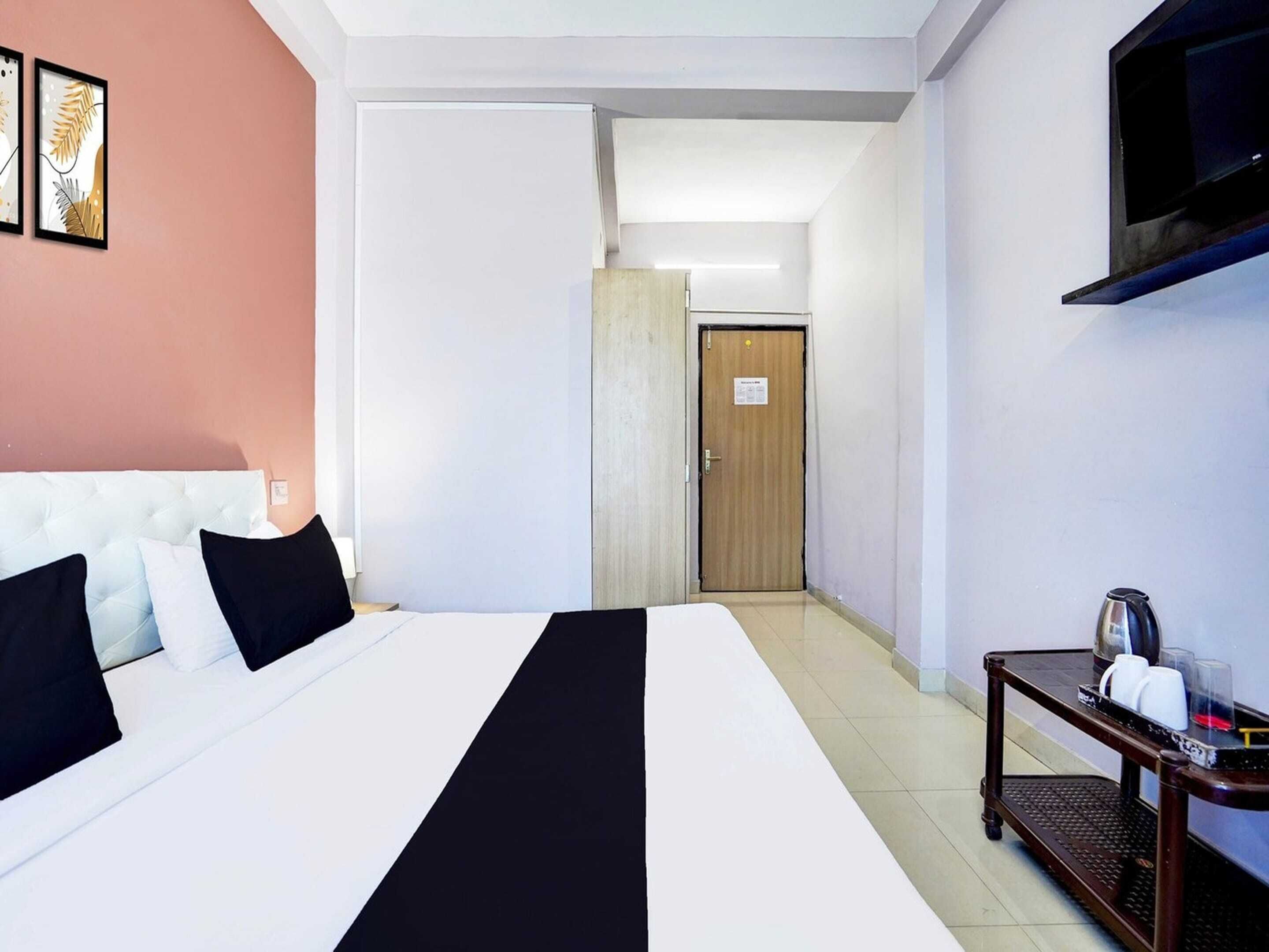 Een hotelkamer met een bed, een nachtkastje met een waterkoker en glazen, een aan de muur bevestigde televisie en een deur naar een andere kamer.