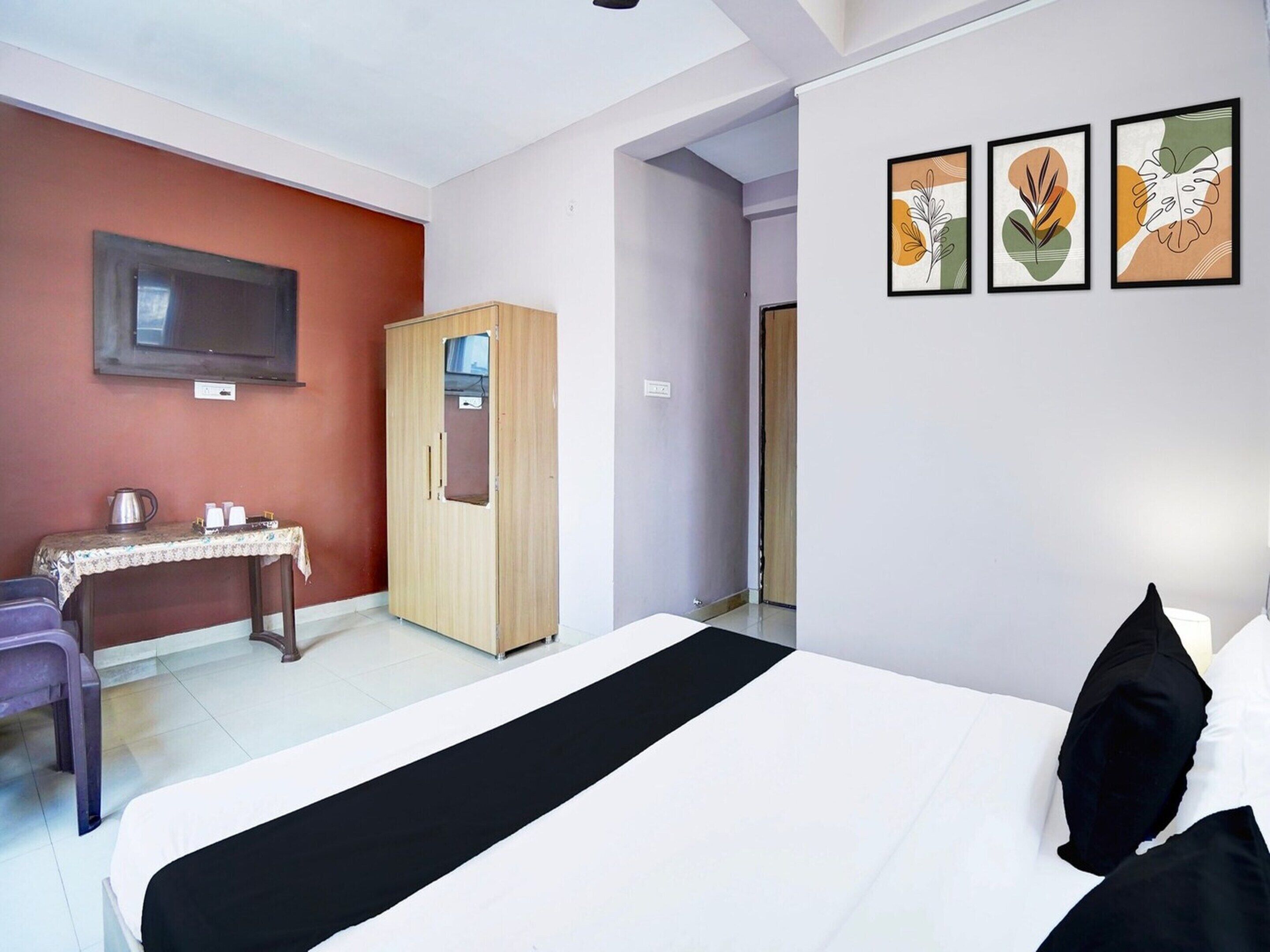 A hotel room with a bed, a desk with a chair, a wardrobe, and three framed artworks on the wall.