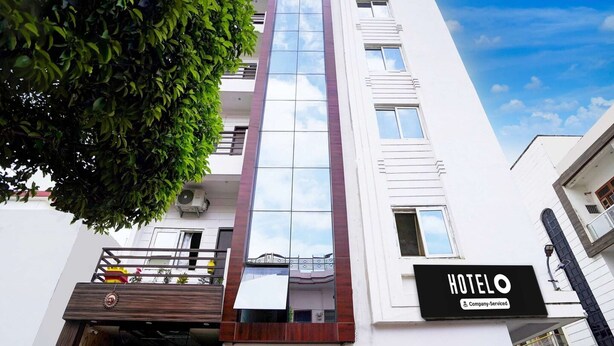 A modern hotel building with a prominent glass facade, a sign indicating it's a company-serviced hotel, and a visible balcony with railings. - Classic With Balcony Hotel SKR (Varanasi)