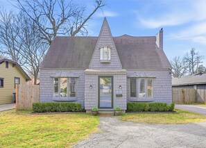 Een twee verdiepingen tellend huis met een puntige dak, een veranda en een kleine voortuin met een grindpad.