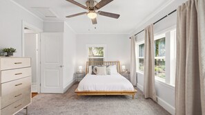 Een slaapkamer met een bed, een plafondventilator, een dressoir, een raam met gordijnen en een nachtkastje met een lamp.