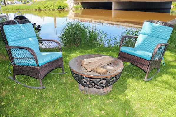 Two wicker rocking chairs with blue cushions, a metal fire pit with logs, and a pond in the background. - Boat or WaveRunner Rentals available next door! Relax or Explore Lake Winnebago! (Fond du Lac)