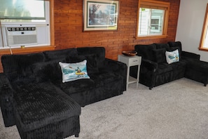 A living room with a brick wall, two black sofas with cushions, a small white side table, and a window with blinds.