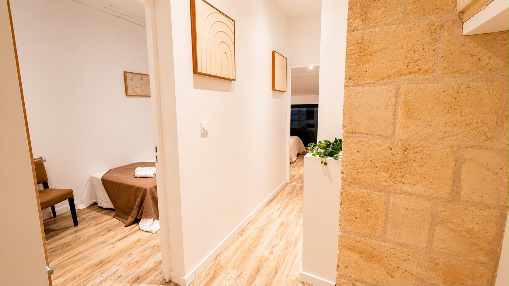 Une chambre d’hôtel avec un plancher en bois, un lit recouvert d’une couverture blanche, une chaise et une petite table. Un mur en briques se trouve d’un côté et un mur blanc de l’autre.