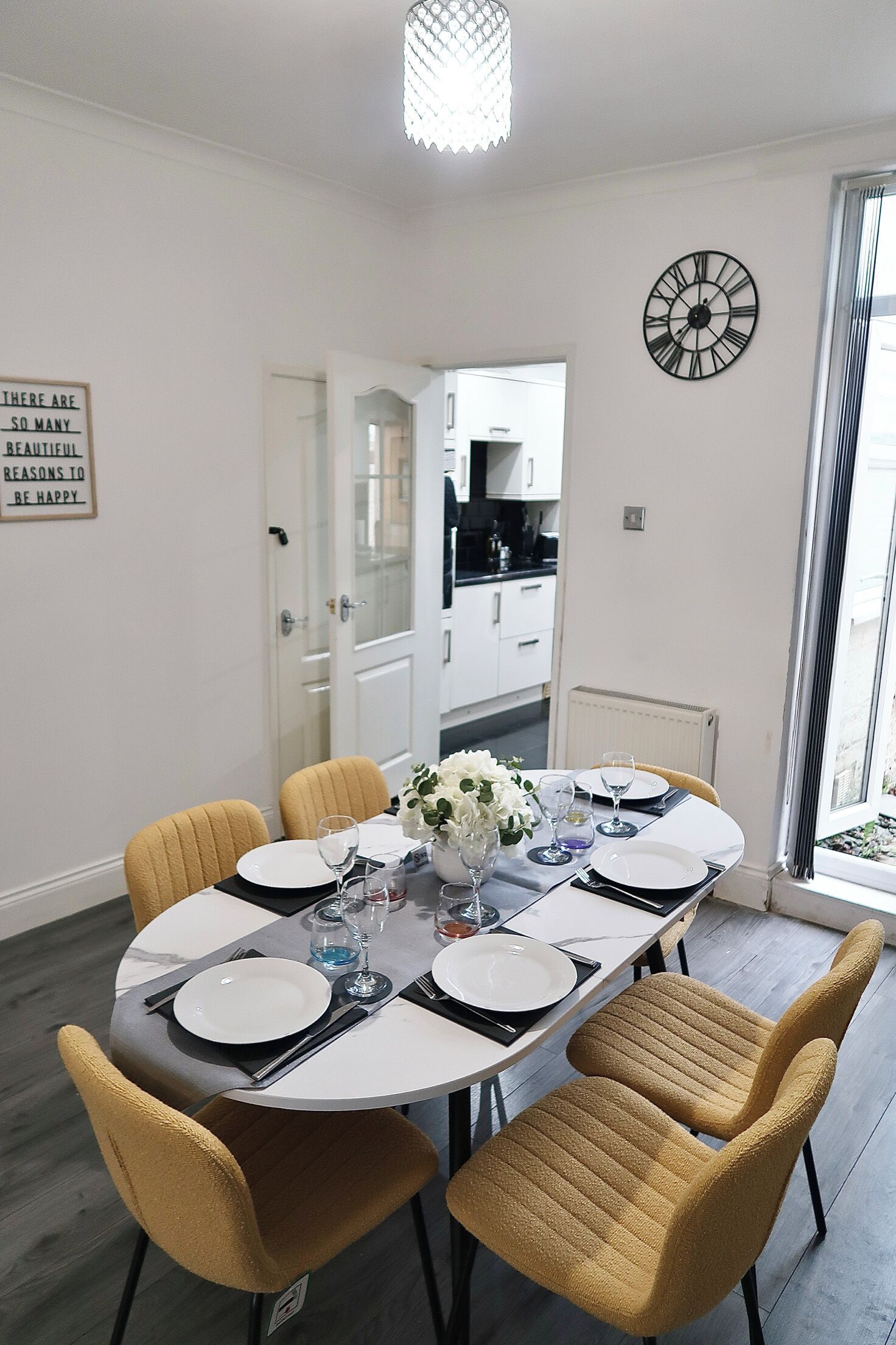 A dining area with a round table set for four, chairs, a clock on the wall, and a kitchen in the background.