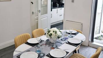 A dining area with a round table set for four, chairs, a clock on the wall, and a kitchen in the background.