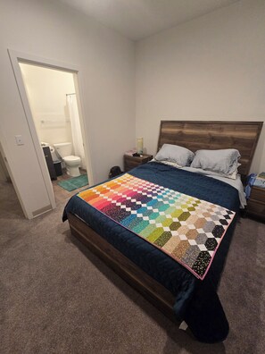 Ein Schlafzimmer mit Bett, Nachttisch, Holz-Kopfstütze und einem durchgehenden Türrahmen sichtbarem Badezimmer.