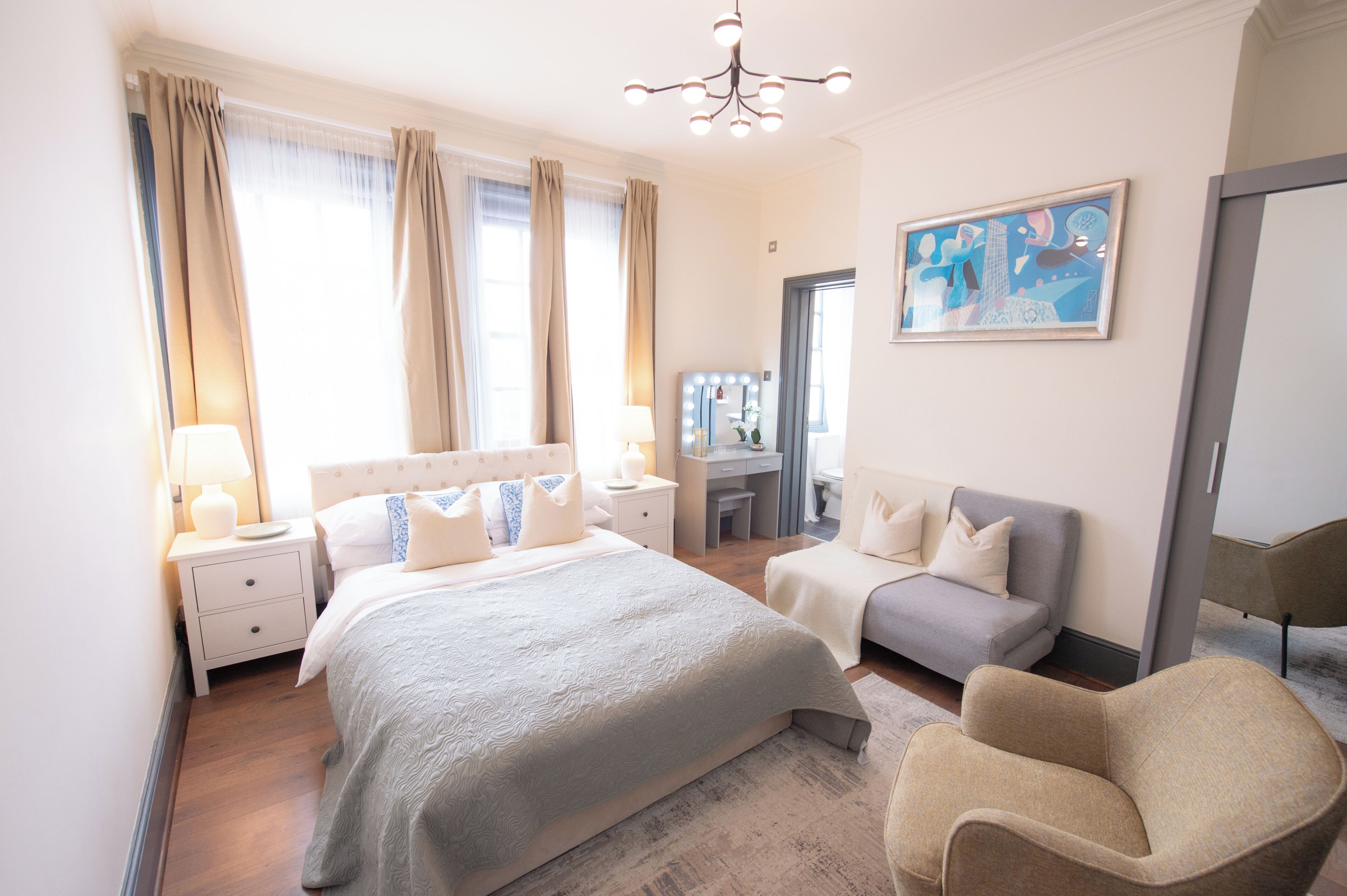 A bedroom with a bed, bedside tables, a desk, a chair, a couch, and a painting on the wall.