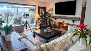 En stue med sofa, sofabord og pejs. Der er et fjernsyn monteret på væggen og en balkon med udendørs møbler.