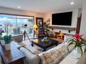 Ein Wohnzimmer mit einer Couch, einem Couchtisch und einem Kamin. Ein Fernseher ist an der Wand montiert und es gibt einen Balkon mit Gartenmöbeln.