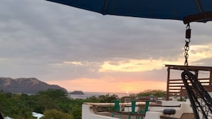 Een balkon met een hangstoel, uitzicht op de zonsondergang en een berg in de verte.