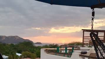 Un balcón con una hamaca colgante, vista del atardecer y una montaña distante.