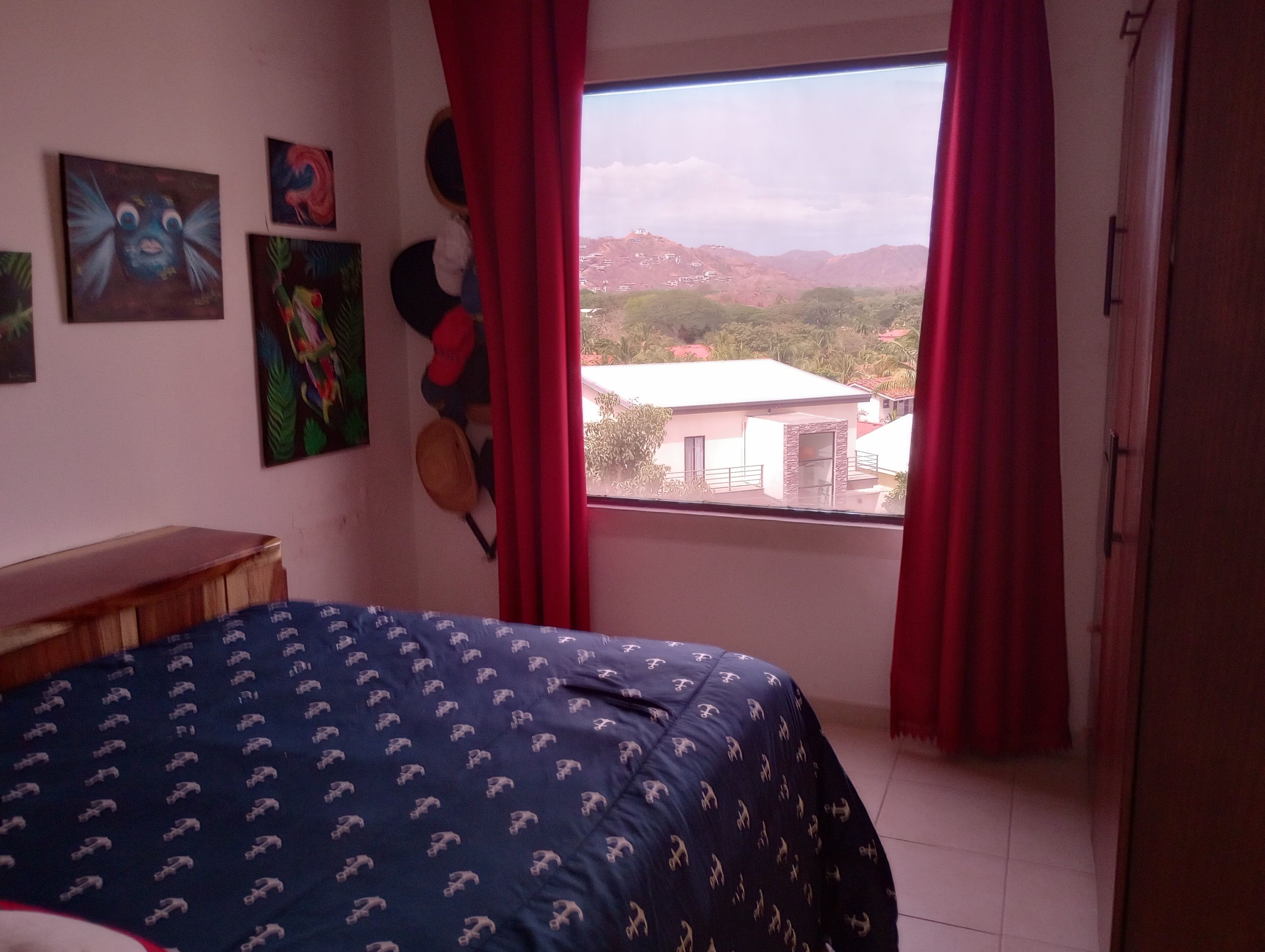 A bedroom with a bed, a window with red curtains, and a view of buildings and hills.
