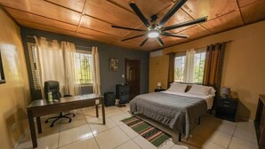 Un dormitorio con cama, escritorio, silla, ventilador de techo y ventana con cortinas.
