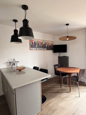 A modern kitchen with a white island, black pendant lights, a wooden dining table, and a TV mounted on the wall.