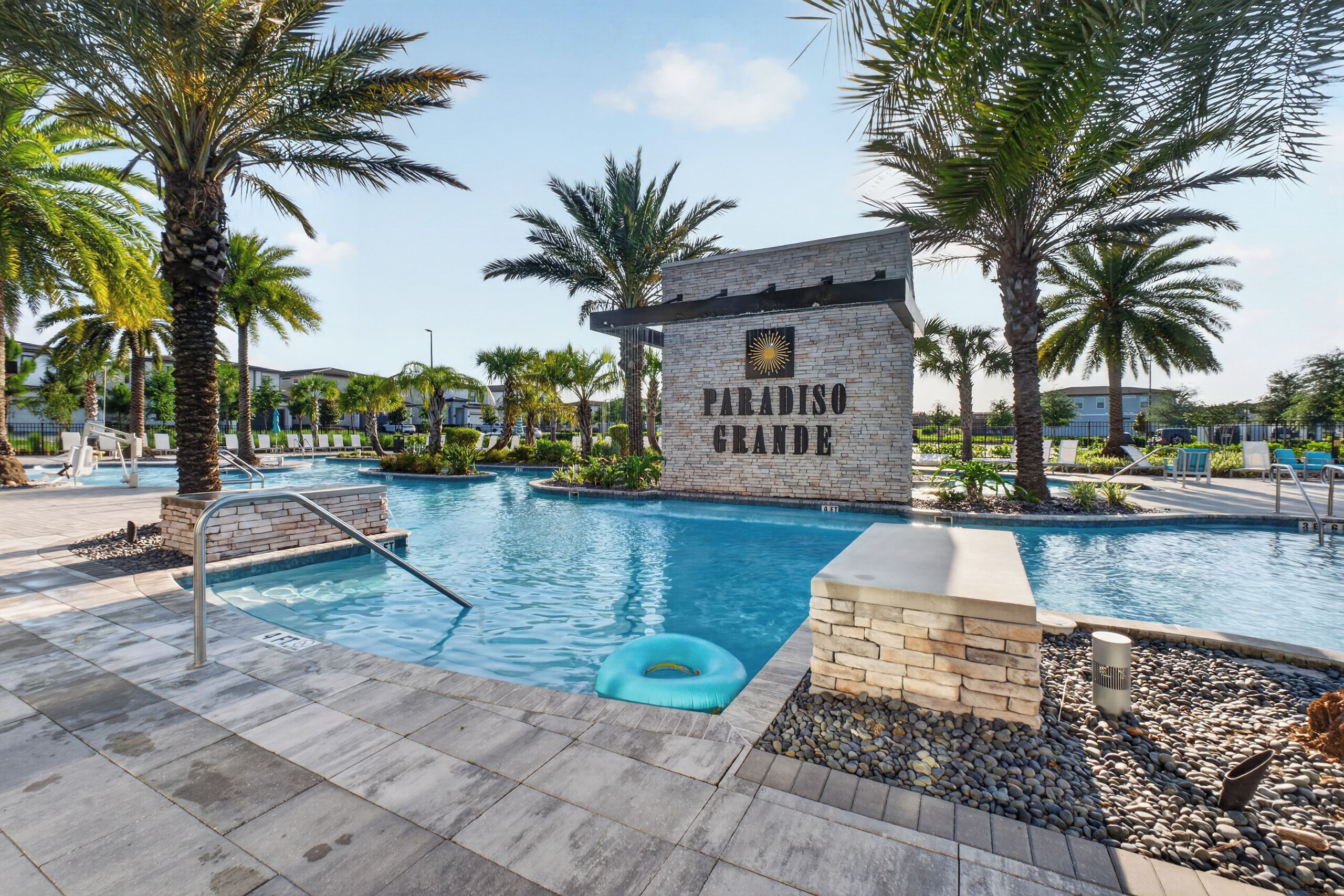A swimming pool area with a sign reading 'PARADISO GRANDE,' surrounded by palm trees and a clear sky.
