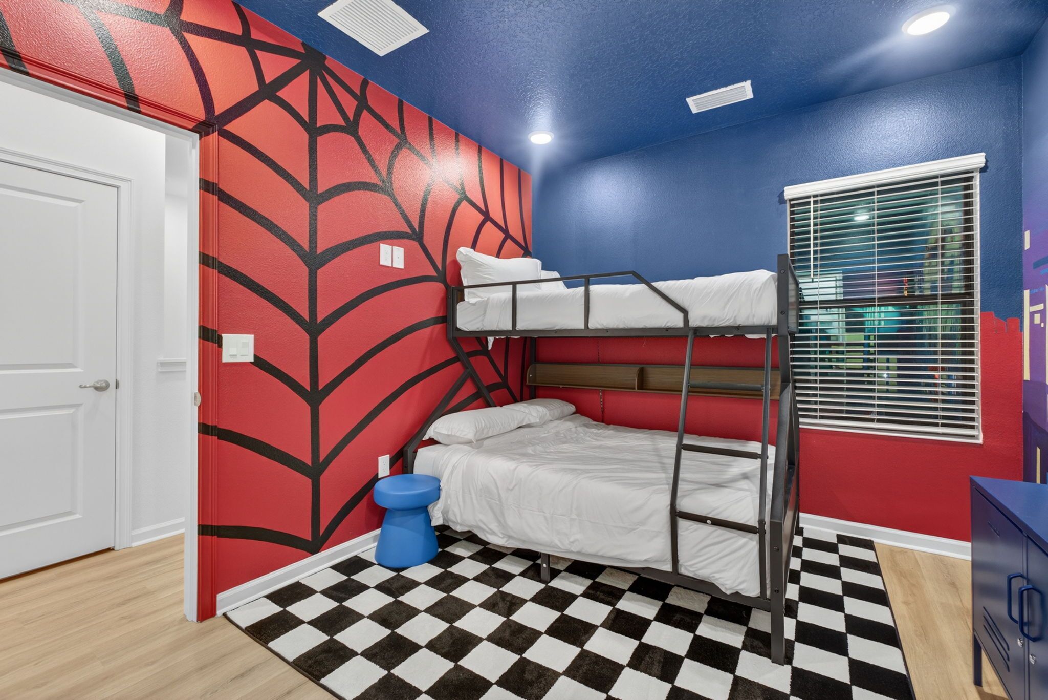 Una habitación con literas, piso a cuadros, pared roja con diseño de telaraña, un taburete azul y una ventana con persianas.