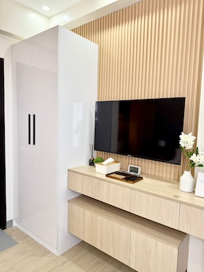 Ein modernes Zimmer mit an der Holzwand montiertem Fernseher, einem weißen Kleiderschrank und einem Holztisch mit Pflanze und Uhr.