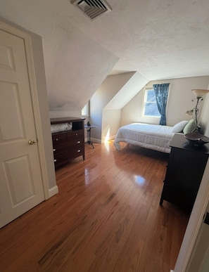 A bedroom with a sloped ceiling, a bed, a dresser, a lamp, and a window with curtains.