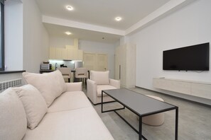 Uma sala de estar moderna com sofá, mesa de centro, TV fixada na parede e cozinha ao fundo.